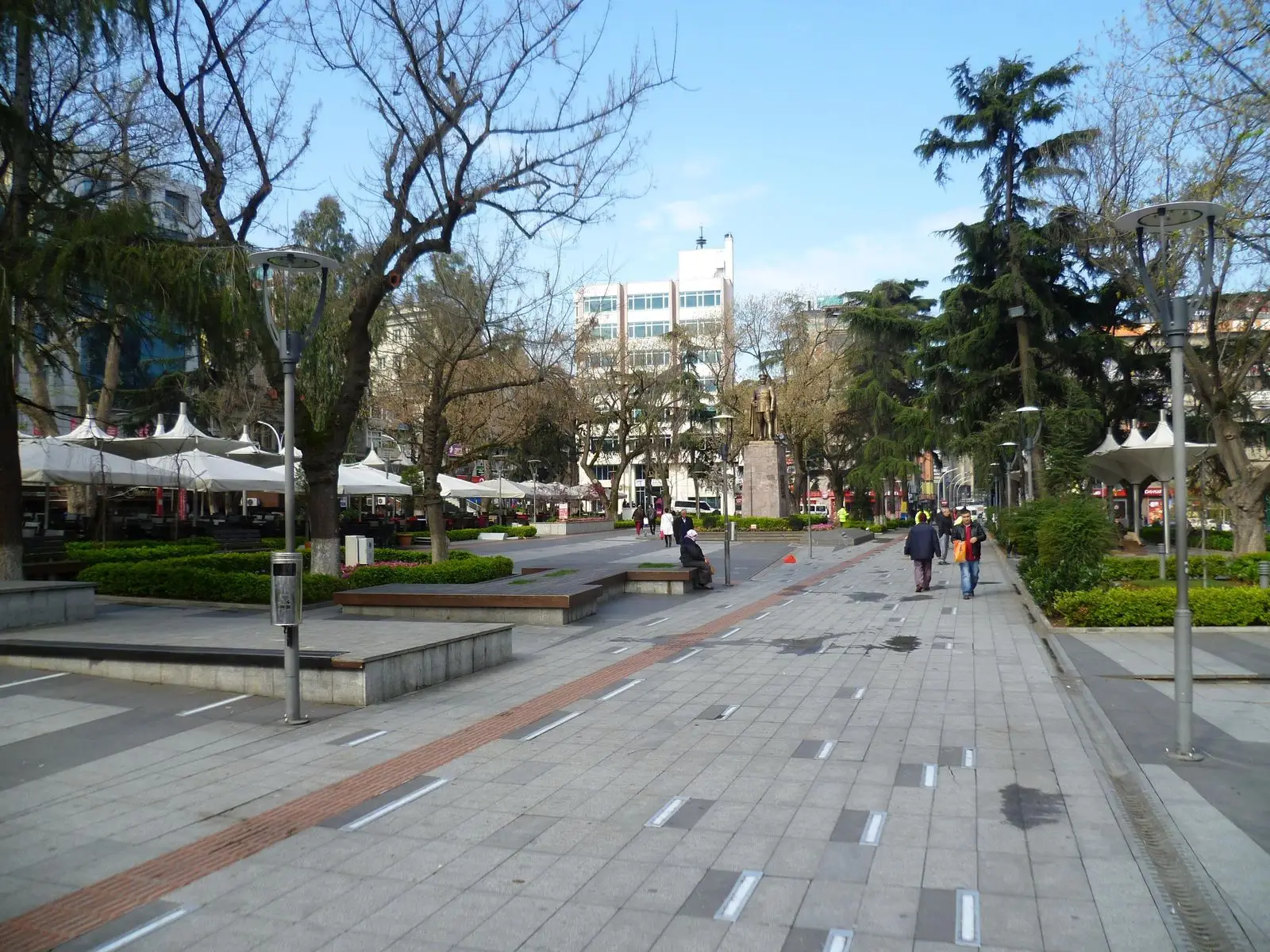 Meydan Parkı: taş döşeli yaya yolu, çınar ve sedir ağaçları, arkada şehir